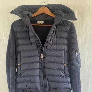 Moncler cardigan - Snygg mörkblå cardigan från Moncler med stickade ärmar och huva. Jackan har dragkedja framtill, två fickor med knappar och en ärmficka med dragkedja. Perfekt för kyliga kvällar. Strlk s. Priset är inte hugget i sten. Inga byten.
