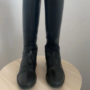 Svarta höga ridstövlar - Säljer ett par klassiska svarta ridstövlar i skinn med rund tå och dragkedja baktill. Stövlarna har diskreta detaljer med knappar vid ankeln och är perfekta för ridning. Storlek 36, använda i ca 1 år, inga repor eller skador ser nya ut. Köpta för 1200kr 