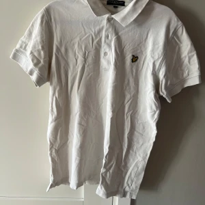 Vit piké från Lyle & Scott - Säljer en klassisk vit pikétröja från Lyle & Scott med korta ärmar och knappslå framtill. Tröjan har det ikoniska gula logobroderiet på bröstet och är tillverkad i bomull. Perfekt för en stilren och sportig look. Använd-i begangnad skick.