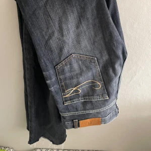 Mörkgrå jeansbyxor med broderad bakficka - Säljer ett par mörkgrå jeansbyxor med snygg gul broderad detalj på bakfickan. Jeansen har raka ben och en klassisk femficksdesign. Perfekta för dig som gillar en stilren men ändå unik look.