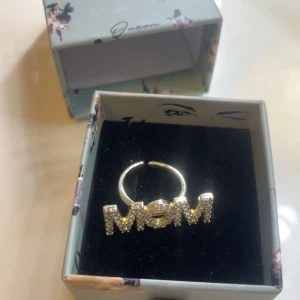 Guldfärgad ring med texten MOM - Trendig guldfärgad ring med texten 'MOM' dekorerad med glittrande stenar. Levereras i en fin ask. Ny pris 599 säljer för 400 och den är ny