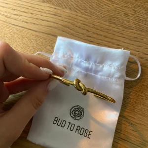 Guldfärgat armband med knut från Bud to Rose - ROSTFRITT STÅL MED 14K GULDPLÄTERING! Stilrent armband i guldfärg från Bud to Rose med en dekorativ knutdetalj framtill. Armbandet är stelt och öppet, vilket gör det enkelt att ta på och av. Perfekt accessoar för att lyfta din outfit. ALDRIG ANVÄNT!!