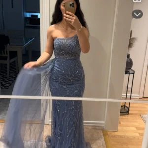 Glittrig blå långklänning med släp - Säljer en glittrig blå långklänning med axelbandslös design och elegant släp. Klänningen har ett vackert mönster av paljetter och pärlor som ger en lyxig känsla. Perfekt för bal eller fest. Figurnära passform och transparent tygdetalj vid sidan.