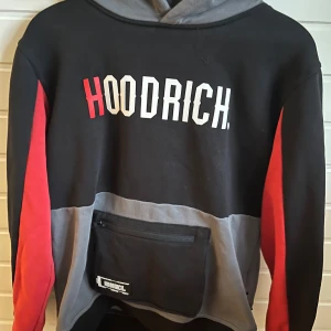 Svart och grå hoodie från Hoodrich - Säljer en snygg hoodie från Hoodrich i svart, grått och rött. Tröjan har stor magficka med dragkedja och tryckt logga på bröstet. Perfekt för en streetwear-look och passar dig som gillar bekväma och stilrena plagg.
