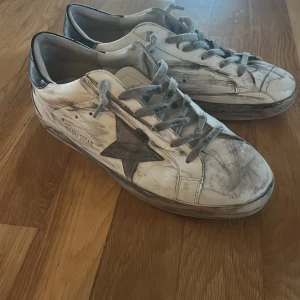 Vita sneakers från Golden Goose med svart stjärna - Golden Goose sneakers i vitt skinn med grå snörning och svart stjärndetalj på sidan. Skorna har svart hälparti och en rund tå. Klassisk modell med platt sula och slitna detaljer för en cool look. Skorna behövs bara tvättas får med kartong och påse 