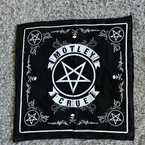 Svart bandana med Mötley Crüe-tryck - Säljer en svart bandana med vitt tryck från Mötley Crüe. Halsduken har pentagram, dödskallar och dekorativa detaljer i hörnen. Perfekt accessoar för dig som gillar rock och vill sticka ut. Helt ny. Bomull. Mått  51 x 51 cm.