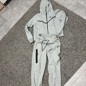 Grå zip-hoodie från Nike och grå Nike byxor  - Säljer en grå zip-hoodie från Nike med svart dragkedja och huva. Jackan har en sportig look med diskret Nike-logga på bröstet och ribbade muddar vid ärmslut. Perfekt för träning eller chill. Med matchande byxor till. 