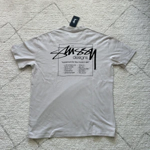 Beige t-shirt från Stüssy med tryck - Säljer en beige t-shirt från Stüssy med svart logga på bröstet och stort tryck på ryggen. T-shirten har rund halsringning och är i mjuk bomull. Perfekt för en avslappnad streetwear-look.