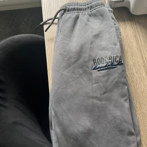 Grå mjukisbyxor från Hoodrich - Grå mjukisbyxor från Hoodrich med resår och dragsko i midjan. Byxorna har sidofickor och en broderad logga på ena benet. Perfekta för chill dagar eller när du vill vara bekväm.