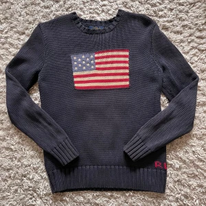 Mörkblå stickad tröja med amerikansk flagga från Polo Ralph Lauren - Säljer en mörkblå stickad tröja från Polo Ralph Lauren med en stor amerikansk flagga på bröstet. Tröjan har rund halsringning och långa ärmar. Klassisk och stilren design med ribbade muddar och nederkant.
