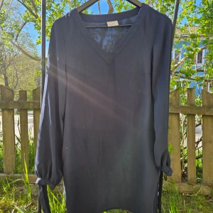 Svart långärmad klänning med v-ringning - En stilren svart klänning med v-ringad hals och långa ärmar. Ärmarna har knytband vid handlederna för en extra detalj. Perfekt för dig som gillar enkel men snygg design. Genomskinligt tyg i ärmarna. 