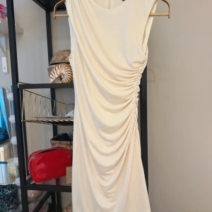 Vit omlottklänning med drapering från Zara - Säljer en elegant vit klänning från Zara i storlek XS. Klänningen har en snygg drapering längs sidan, är ärmlös och har omlottdetalj samt dragkedja i ryggen. Perfekt för dig som gillar stilrena och moderna plagg. Passar även mig som är en storlek S! 