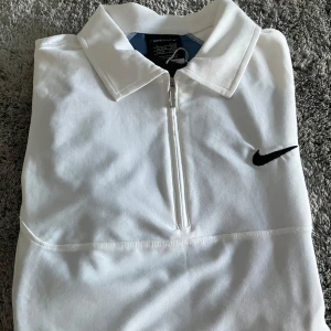 En NY golftröja med dragkedja från Nike - Snygg vit golftröja från Nike med halv dragkedja och svart logga på bröstet. Tröjan har korta ärmar, krage och ventilerande meshpanel på ryggen för extra komfort. Perfekt för golfbanan eller träning. 88% polyester, 12% elastan. Storlek S, men väldigt stor i storleken! Aldrig använd pga storleken. 