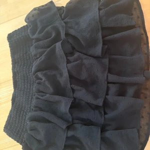 Svart volangkjol med prickigt mönster - Supersöt svart kjol med flera lager volanger och diskret prickigt mönster. Kjolen är i storlek 146/152, men skulle säga att damen passar som en XS. Kjolen har bred, smockad midja och är gjord i ett lätt, transparent tyg som ger en luftig känsla. Perfekt för dig som gillar en feminin och trendig stil. ALLTID 10% RABATT NÄR DU KÖPER 2 PLAGG ELLER FLER💗🫶!!!