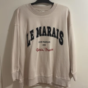 Beige sweatshirt med tryck 'Le Marais' - Säljer en oversized beige sweatshirt med texten 'Le Marais Club Français 1995 Paris, France' framtill. Tröjan har rund halsringning och långa ärmar med breda muddar. Perfekt för en avslappnad stil.