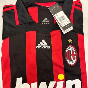 AC Milan fotbollströja från Adidas - Alla storlekar finns för leverans inom 3-4 dagar Säljer en klassisk AC Milan fotbollströja från Adidas med röda och svarta vertikala ränder, klubbmärke och stjärna på bröstet samt vit Bwin-logga. Tröjan har korta ärmar och gulddetaljer längs sidorna. Perfekt för fans!