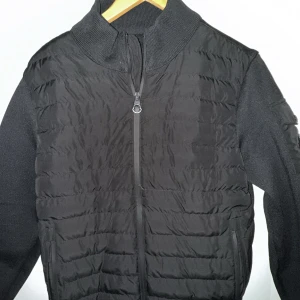 Svart quiltad cardigan jacka från Moncler - Snygg svart jacka från Moncler med quiltad framsida och stickade ärmar. Jackan har hög krage, dragkedja framtill och Moncler-logga på bröstet. Perfekt för dig som vill ha en stilren och bekväm jacka med exklusiv känsla.