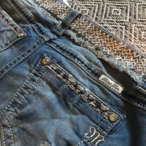Blå Miss Me jeans capri med mönstrad midja - Säljer ett par skitcoola blå Miss Me jeans capri med snygga slitningar och dekorativa detaljer i midjan. Byxorna har fransig kant nertill, broderade detaljer och silverfärgade knappar. Perfekt för dig som gillar unika jeans med extra detaljer.