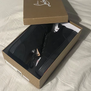 Svarta sneakers från Christian Louboutin - Säljer ett par svarta sneakers från Christian Louboutin med klassisk röd sula. Skorna har snörning, rund tå och är tillverkade i mocka. Insidan är beige och de har en stilren, minimalistisk design som passar till många outfits.