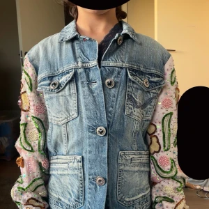Jeansjacka med broderade ärmar från Replay - Unik jeansjacka från Replay med ljusblå tvätt och broderade ärmar i rosa tyg. Ärmarna har blommiga mönster, pärlor och paljetter i grönt, gult och rosa. Klassisk krage, knappar framtill och fyra fickor. Perfekt statement-plagg för dig som vill sticka ut.