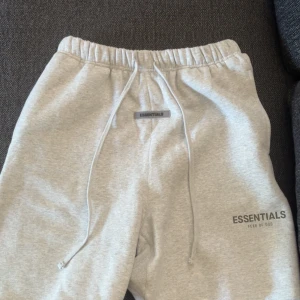  Essentials short - Säljer ett par grå shorts från Essentials Fear of God. Shortsen har resår i midjan med snörning och logga tryckt på benet samt en patch framtill. Perfekta för en avslappnad stil.