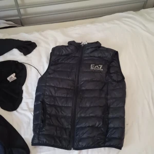 Svart dunväst från Emporio Armani EA7 - Säljer en svart dunväst från Emporio Armani EA7 med dragkedja framtill och hög krage. Västen har ett quiltat mönster och logga på bröstet. Perfekt för lager-på-lager och stilren look.