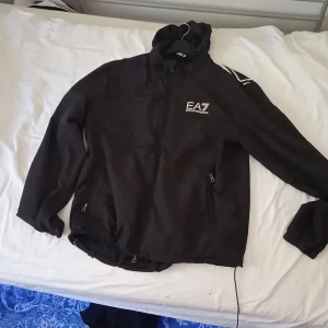 Svart vindjacka från EA7 Emporio Armani - Säljer en svart vindjacka från EA7 Emporio Armani med huva och dragkedja. Jackan har vita logotyper på bröstet och ryggen samt två fickor med dragkedja. Perfekt för dig som gillar stilren och sportig design.