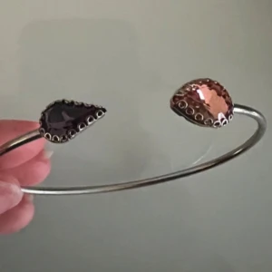 Silverfärgat armband med droppformade smaragder  - Snyggt armband i silverfärg 925 äkta doppad i guld med två droppformade smaragder i lila och rosa. Stenarna sitter i varsin ände av det öppna armbandet och har en dekorativ infattning. Perfekt accessoar för att lyfta din stil. Pris kan diskuteras 