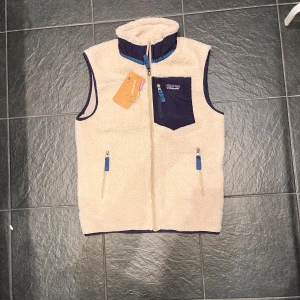 Beige fleeceväst från Patagonia - Säljer en beige fleeceväst från Patagonia med marinblå detaljer och dragkedjor. Västen har en hög krage, två fickor fram och en bröstficka med logga. Perfekt för lager-på-lager och outdoor.