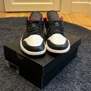 Nike Air Jordan 1 Low Black/Fire Red-White - Säljer ett par Nike Air Jordan 1 Low i svart, vitt och rött. Skorna har klassisk låg siluett, svart ovandel med vita och röda detaljer samt perforerad tå. Röd insida och broderad logga på hälen. Perfekta för dig som gillar streetwear.