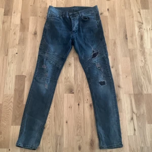 Mörkblå slitna jeans med ribbade detaljer - Säljer ett par mörkblå jeans med skinny fit och ribbade detaljer över knäna. Jeansen har flera slitningar och hål framtill för en tuff look. Klassisk femficksmodell och knappgylf.