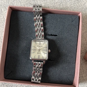 Silverfärgad rektangulär klocka från Yazole - Stilren klocka från Yazole med rektangulär urtavla och länkarmband i silverfärgad metall. Urtavlan är vit med diskreta stenar som timmarkeringar och har en klassisk, elegant look. Perfekt accessoar för att lyfta din outfit. Den är helt ny och aldrig använd. Säljer pågrund av att jag är guldtjej. Pris går att diskutera. 