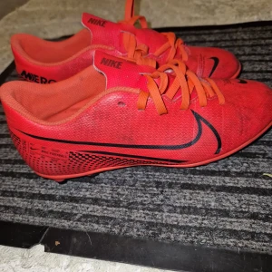 Röda Nike Mercurial fotbollsskor - Säljer ett par röda Nike Mercurial fotbollsskor med svarta detaljer och snörning. Skorna har rund tå och platt sula med dobbar, perfekt för spel på gräs. Snygg design med stor svart Nike-logga på sidan.