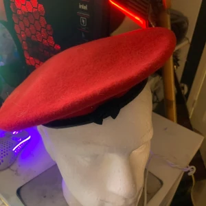 Röd basker från Kangol - Säljer en klassisk röd basker från Kangol i 100% ull. Den har en rund form och är dekorerad med en liten svart rosett på insidan. Perfekt accessoar för att lyfta din stil med en färgklick.
