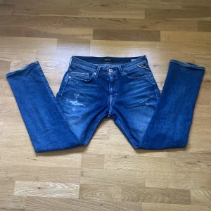 Blåa replay jeans  - Säljer ett par blå Replay Mini Waitom jeans med slitna detaljer och klassisk femficksdesign. Jeansen har normal passform och raka ben. Märkeslapp bak i midjan och Replay-logga vid fickan. Perfekt för en person som vill ha enavslappnad stil.