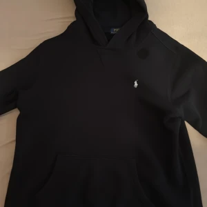 Svart hoodie från Polo Ralph Lauren - Säljer en svart hoodie från Polo Ralph Lauren med klassisk broderad logga på bröstet. Tröjan har huva och en stor magficka framtill. Perfekt för en chill och stilren look.