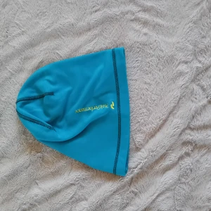 Turkos mössa från Peak Performance - Turkos mössa från Peak Performance med gul logotyptryck på sidan. Tillverkad i ett tunt och stretchigt material, perfekt för träning eller utomhusaktiviteter. Snygga kontrastsömmar och liten Signatur PeakPerformance R&D-tag på kanten.