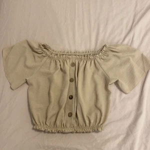 Beige croppad blus med knappar - Säljer en söt beige croppad blus med korta ärmar och resår vid halsringning och midja. Blusen har dekorativa knappar framtill och en luftig passform, perfekt för varmare dagar.