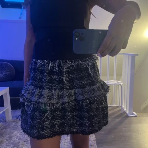 Svart och grå rutig kjol med volang - Kort kjol i svart och grått med rutigt mönster. Kjolen har en volangdetalj. Storlek Xs