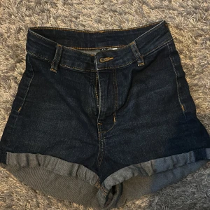 Mörkblå jeansshorts med uppvik - Säljer ett par mörkblå jeansshorts med klassisk femficksmodell och uppvikta benslut. Shortsen har ganska hög midja och synliga sömmar i kontrastfärg. Perfekta för varma dagar och enkel att matcha med olika toppar.