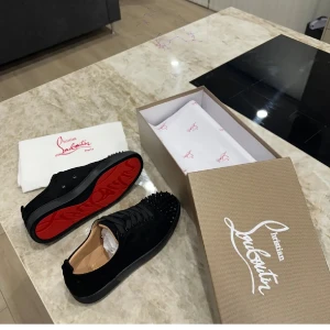 Christian Louboutin - Säljer ett par svarta sneakers från Christian Louboutin med ikoniska röda sulor och svarta nitar på ovandelen. Skorna har snörning och är tillverkade i mocka med detaljer i skinn. Perfekta för dig som vill sticka ut med en lyxig och edgy stil.