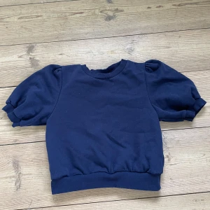 Mörkblå tröja med puffärmar från Gina Tricot - Säljer en mörkblå tröja från Gina Tricot med korta puffärmar och ribbade muddar vid hals, ärmslut och nederkant. Tröjan har en enkel och stilren design som passar perfekt till vardags.