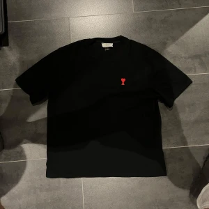 Svart t-shirt från Ami Paris med röd broderad logga - Svart t-shirt från Ami Paris med en liten röd broderad logga på bröstet. Klassisk passform och rund halsringning. Perfekt för en stilren och enkel look.