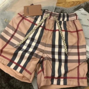 Rutig badshorts från Burberry - Snygga badshorts från Burberry i klassiskt rutig design med beige, svart, vit och röd färg. Shortsen har snörning i midjan och en bakficka med knapp. Perfekta för sommaren och strandhäng.