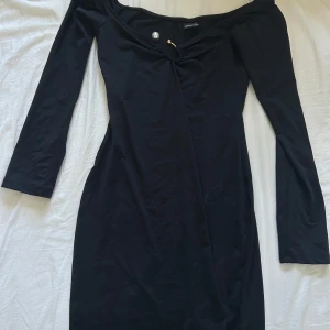 Kort klänning - Säljer en svart  klänning med offshoulder-design. Klänningen har en dekorativ detalj vid halsringningen med en pärla och guldfärgad metall. Tajt passform och kort. Aldrig använd !!