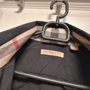 Mörkblå skjorta från Burberry  - Säljer en klassisk mörkblå skjorta från Burberry Brit med ikoniskt rutigt tyg på insidan av krage och manschetter. Skjortan har knappar framtill och lång ärm. Perfekt för dig som gillar stilrena plagg med en twist.