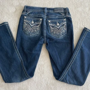 Mörkblå jeans med broderade fickor från Miss Me - Snygga mörkblå jeans från Miss Me med unika broderade detaljer på bakfickorna. Jeansen har en låg/medel midja och en bootcut-fit. Köpte dem I somras och de är nästan helt oanvända I superbra skick! Mått: innerbenslängd 81 cm, ytterbenslängd 105 cm. Skriv för fler bilder.
