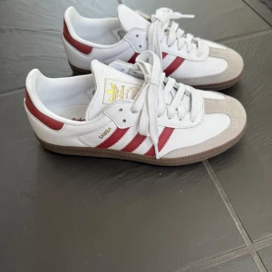 Adidas Samba med röda ränder - Säljer ett par klassiska Adidas Samba sneakers i vitt skinn med röda ränder och detaljer. De är endast använda två gånger så dom är i otroligt bra skick som ni kan se på bilderna.