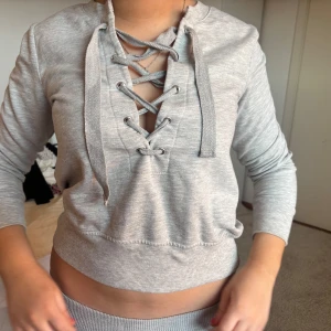 Grå tröja med snörning från Gina Tricot - Säljer en ljusgrå tröja från Gina Tricot med snygg snörning framtill och trekvartsärm. Tröjan har ribbade muddar och är i mjukt material, perfekt för en avslappnad stil.