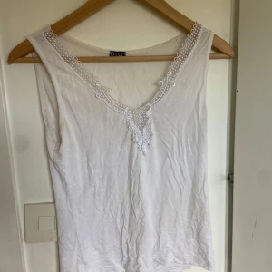 Vit linnetopp med spets från Dolce & Gabbana - Säljer en vit linnetopp från Dolce & Gabbana med v-ringning och fina spetsdetaljer längs halsringningen. Toppen är ärmlös och tillverkad i mjuk bomull, perfekt för varma dagar.